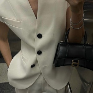 May include: Cream-colored vest with black buttons and a matching pair of trousers. A small black handbag with a gold-colored clasp is held in the hand. A silver bracelet is also visible.