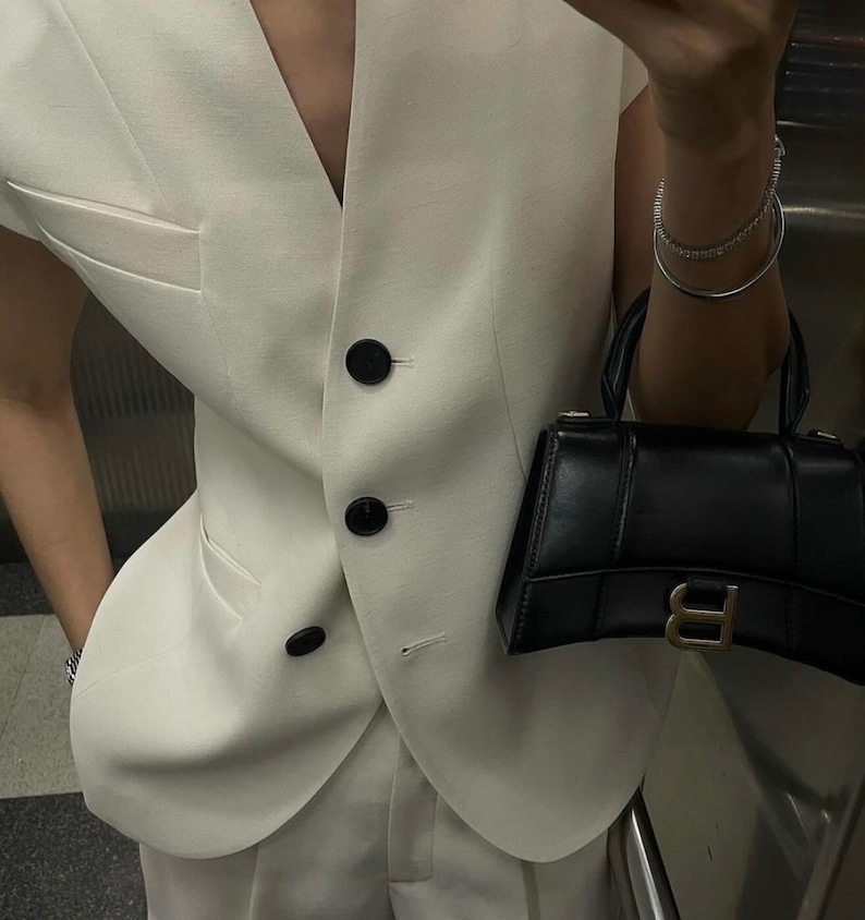May include: Cream-colored vest with black buttons and a matching pair of trousers. A small black handbag with a gold-colored clasp is held in the hand. A silver bracelet is also visible.