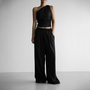 Pu&ograve; includere: Un top nero a spalla unica con un dettaglio drappeggiato sul seno, abbinato a pantaloni neri a gamba larga.