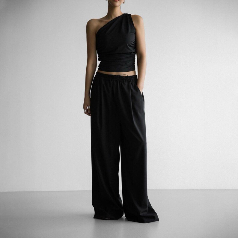 Pu&ograve; includere: Un top nero a spalla unica con un dettaglio drappeggiato sul seno, abbinato a pantaloni neri a gamba larga.