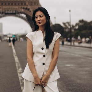 May include: A woman in a cream-colored suit stands in front of the Eiffel Tower. The suit includes a sleeveless jacket with black buttons and wide-leg pants. She carries a black purse with a gold chain strap.