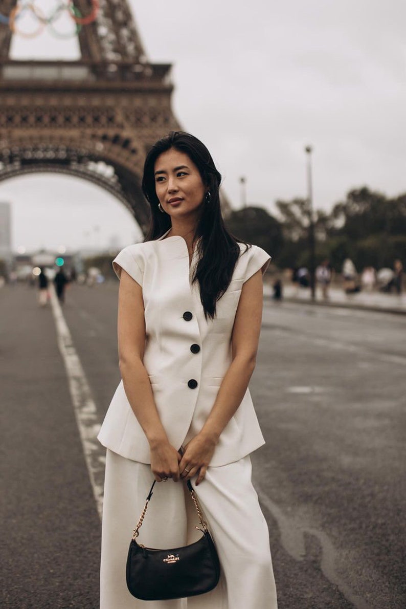 May include: A woman in a cream-colored suit stands in front of the Eiffel Tower. The suit includes a sleeveless jacket with black buttons and wide-leg pants. She carries a black purse with a gold chain strap.