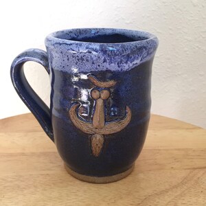 May include: A blue ceramic mug with a brown, stylised design of a face with a moustache. The mug has a white glaze on the rim and a light blue glaze on the inside.