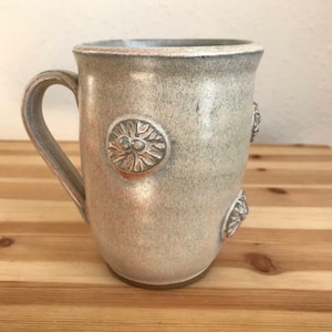 Puede incluir: Una taza de cerámica blanca con una superficie texturizada y tres diseños florales en relieve. La taza tiene un asa y una forma redondeada.