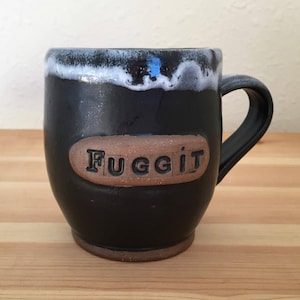 May include: Black ceramic mug with a white rim and a brown label that says "FUGGIT".