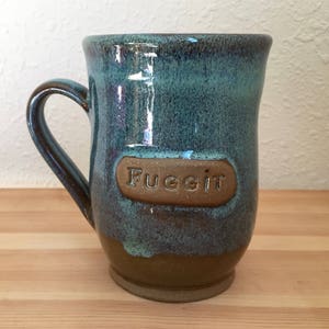 May include: A blue and brown ceramic mug with a brown label that says "Fuggit".