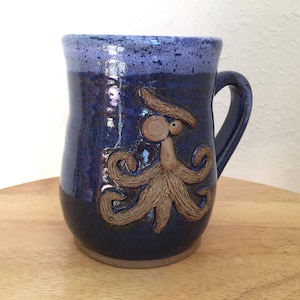 May include: A blue ceramic mug featuring a brown octopus design. The octopus has a face with two eyes and tentacles.
