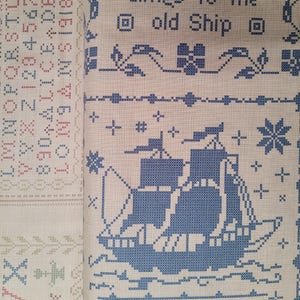 May include: Cross-stitch sampler with a blue ship design and the text "clings to the old Ship." The sampler also features decorative borders, floral motifs, and an alphabet. The background is a light beige color.