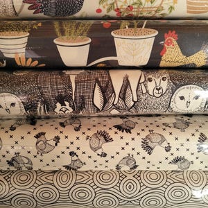 May include: A stack of decorative fabric rolls featuring various patterns. Designs include chickens, potted plants, and geometric shapes in black, white, and shades of brown. The fabrics appear to be cotton or a similar material.
