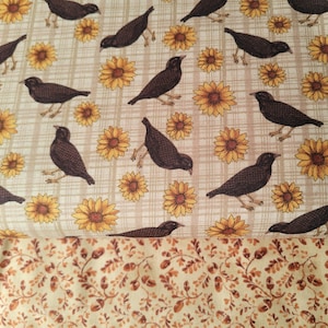 May include: A fabric print featuring a pattern of black birds and yellow sunflowers on a beige plaid background, layered over a second fabric section with a dense, repeating pattern of small brown acorns and leaves on a light cream background.