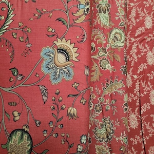 May include: Close-up of red fabric with a floral pattern. The design features large, stylized flowers in shades of blue, tan, and green, with intricate details. The fabric is folded, revealing a similar pattern in a lighter shade.