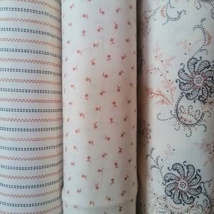 May include: Three fabric rolls with different patterns. The first roll has thin white and orange stripes. The second roll has a white background with small orange floral designs. The third roll has a white background with a black and orange paisley pattern.