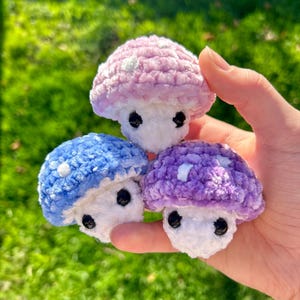May include: Three handmade crochet mushroom ornaments in shades of pink, blue, and purple. Each mushroom has a white stem and small black eyes. The caps feature white polka dots. The mushrooms are held in a hand against a blurred green background.