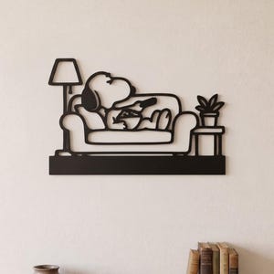 Könnte beinhalten: Ein schwarzes Wandkunstobjekt aus Metall, das einen Cartoon-Hund zeigt, der auf einem Sofa liegt und eine Fernbedienung hält. Links steht eine Stehlampe, rechts ein kleiner Beistelltisch mit einer Topfpflanze. Das Design ruht auf einem festen Sockel.