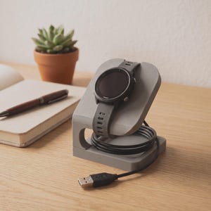 Peut inclure: Un support de charge gris imprimé en 3D pour montre connectée, doté d'une structure de support incurvée et d'une découpe circulaire à la base pour le câble. Une montre noire avec un bracelet en silicone texturé est posée sur le support.