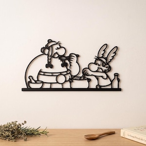 May include: A black metal wall art piece featuring a silhouette outline of the cartoon characters Asterix and Obelix cooking together. The design shows Obelix eating from a spoon while Asterix stirs a pot, with a bowl and bottle nearby.