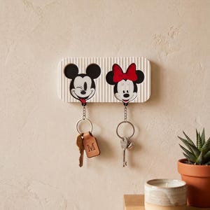Könnte beinhalten: Ein weißer, rechteckiger, gerippter Wand-Schlüsselhalter mit zwei kreisförmigen Aussparungen, die Mickey Mouse mit einem Augenzwinkern und Minnie Mouse mit einer roten Schleife zeigen. Zwei Metallschlüsselanhänger hängen daran, einer mit Lederanhänger.