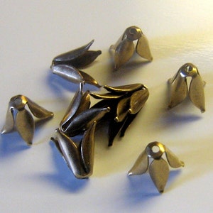 Op de afbeelding: Een groep van acht goudkleurige metalen bloemvormige versieringen. De versieringen hebben vijf bloemblaadjes en een klein gaatje in het midden.