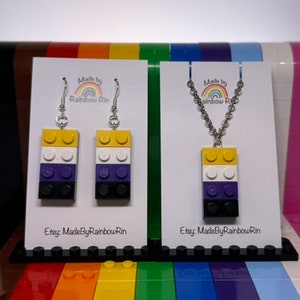 以下が含まれることがあります： レゴブロックで作られたイヤリングとネックレスのペア。イヤリングとネックレスは、ノンバイナリープライドフラッグの色、イエロー、ホワイト、パープル、ブラックです。イヤリングはシルバーのフックにぶら下がっています。ネックレスはシルバーチェーンです。カードには「Made by Rainbow Rin」と「Etsy: MadeByRainbowRin」のテキストが書かれています。
