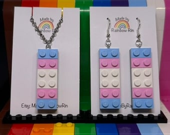 May include: A set of transgender pride jewelry made from LEGO bricks. The set includes a necklace and a pair of earrings. The necklace features a silver chain with a blue, pink, and white LEGO brick pendant. The earrings are silver hooks with blue, pink, and white LEGO brick pendants. The jewelry is displayed on a white background with a rainbow backdrop.