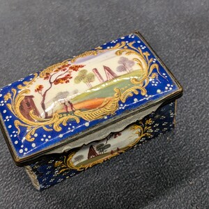 Puede incluir: Una caja rectangular con bisagras que presenta un acabado de esmalte azul intenso con detalles de puntos blancos y volutas doradas. La tapa y el panel frontal muestran paisajes pintados a mano con árboles, edificios y figuras.