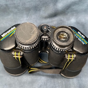 Puede incluir: Binoculares negros con detalles amarillos y azules. Los binoculares tienen un aumento de 10x50 y son de óptica recubierta. El texto "Miranda" está impreso en el lateral de los binoculares.