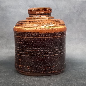 Pode incluir: Um pote de cerâmica marrom e preto com tampa. O pote tem uma superfície texturizada e uma forma ligeiramente arredondada.