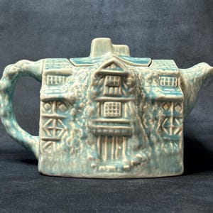 May include: A light blue ceramic teapot shaped like a house. The teapot features detailed architectural elements, including windows, doors, and a textured facade. The handle and spout are integrated into the house design.