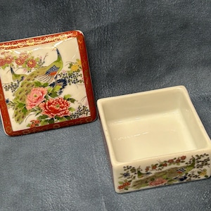 May include: A white porcelain trinket box with a red and gold trim. The lid features a colorful design of peacocks and flowers. The inside of the box is empty.