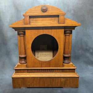 May include: A brown wooden clock case featuring a circular opening, architectural columns on both sides, and a pediment top with a central circular ornament. A small paper label inside reads H. Pontifex & Sons, Makers, London, 506 Oxford Street.