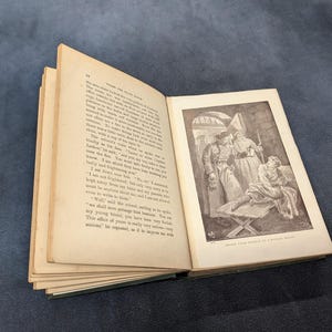 May include: An open antique book with aged, yellowed pages. The right page features a black and white illustration of figures in a room. The book is set against a dark blue background.