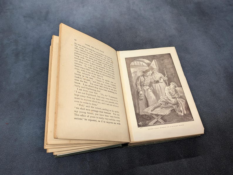 May include: An open antique book with aged, yellowed pages. The right page features a black and white illustration of figures in a room. The book is set against a dark blue background.