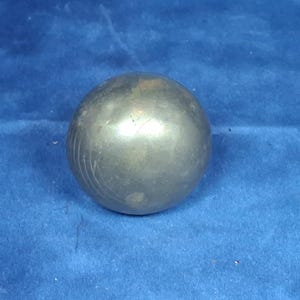 Puede incluir: Un objeto esférico de metal con un acabado de color latón opaco y deslustrado. La superficie de la esfera muestra signos sutiles de desgaste, incluyendo rasguños tenues y una ligera decoloración, con una forma suave y redondeada.