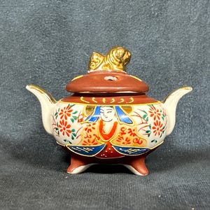 May include: A small, white ceramic incense burner with a red lid and a gold lion finial. The burner is decorated with floral and geometric patterns in red, blue, and gold.