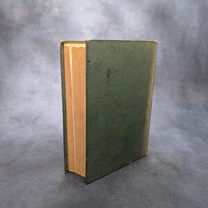 May include: A vintage book with a dark green cover and aged, yellowed pages. The book is standing upright, showcasing the spine and the side of the pages. The cover has a slightly worn appearance, suggesting its age.