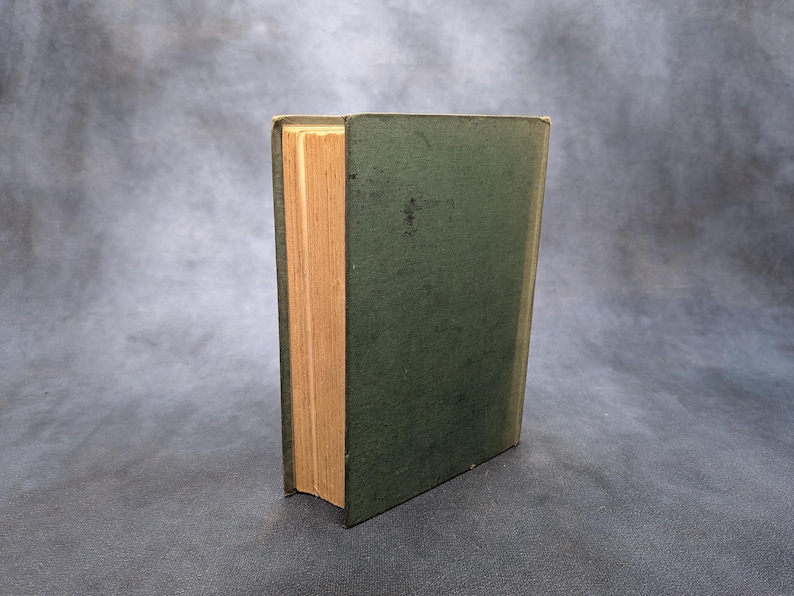 May include: A vintage book with a dark green cover and aged, yellowed pages. The book is standing upright, showcasing the spine and the side of the pages. The cover has a slightly worn appearance, suggesting its age.