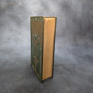 May include: An antique book titled "Under the Jackstaff" with a green cover featuring an illustrated tree design. The book's pages are aged, and the spine is visible. The book is set against a neutral background.