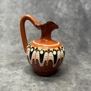 Può includere: Un piccolo brocca in ceramica marrone con un design dipinto in bianco e nero. La brocca ha un corpo rotondo e un beccuccio corto e curvo.