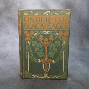 May include: A vintage book titled "UNDER THE BLACK EAGLE" with a green cover. The cover features an ornate design with orange and white accents, including the title text and floral motifs. The book is standing upright.
