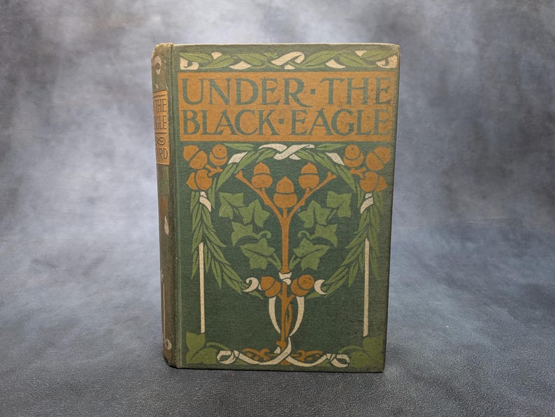 May include: A vintage book titled "UNDER THE BLACK EAGLE" with a green cover. The cover features an ornate design with orange and white accents, including the title text and floral motifs. The book is standing upright.
