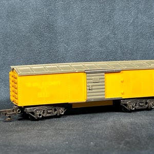May include: A yellow and gray toy train boxcar with black wheels and a side door. The boxcar has a gray roof and a ladder on the side.