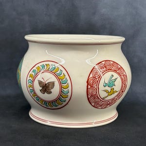 Puede incluir: Un macetero de cerámica blanca con forma redondeada y un borde ancho. El macetero está decorado con tres diseños circulares que presentan una mariposa, pájaros y patrones en espiral en rojo, azul, verde y amarillo.