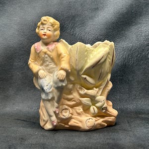 Puede incluir: Un macetero de porcelana con una figura de un joven con una chaqueta amarilla sentado en un tocón de árbol con un diseño floral. El macetero es en tonos de amarillo, blanco y marrón.