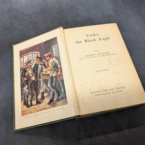 May include: Open vintage book titled "Under the Black Eagle" by Andrew Hilliard. The book's cover features a color illustration of men in military uniforms. The book is illustrated by Blackie and Son Limited.