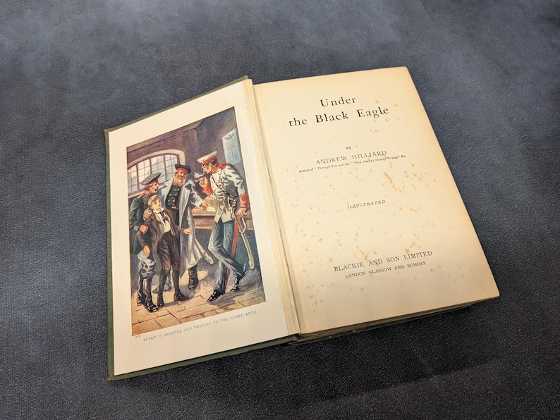 May include: Open vintage book titled "Under the Black Eagle" by Andrew Hilliard. The book's cover features a color illustration of men in military uniforms. The book is illustrated by Blackie and Son Limited.