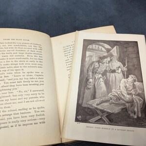 May include: An open book with text and a black and white illustration. The illustration depicts a man in a prison setting, surrounded by figures in coats and hats. The text on the page reads "UNDER THE BLACK EAGLE".