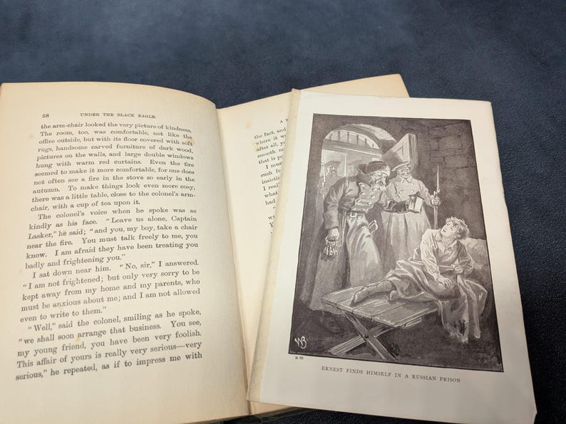 May include: An open book with text and a black and white illustration. The illustration depicts a man in a prison setting, surrounded by figures in coats and hats. The text on the page reads "UNDER THE BLACK EAGLE".