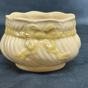 May include: A white ceramic bowl with a yellow ribbon design. The bowl has a scalloped rim and is decorated with a raised yellow ribbon design.