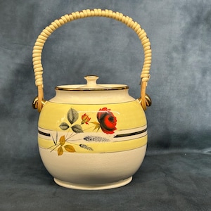 May include: A white ceramic cookie jar with a yellow and white floral design and a woven handle. The jar has a gold trim and a lid.