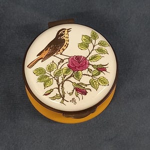 Op de afbeelding: Een ronde, gele sieradenbox met een wit deksel met een handgeschilderd beeld van een bruine en gele vogel die op een rozenstruik met roze rozen zit.